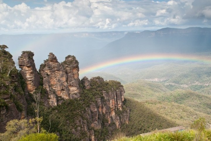 Rock formation with rainbow and distant mountains in a cloudy sky.
