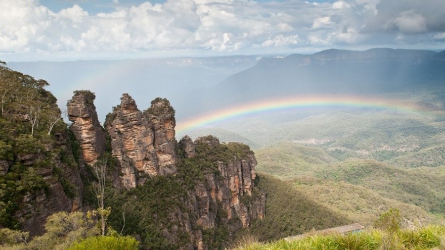 Rock formation with rainbow and distant mountains in a cloudy sky.